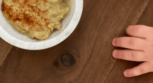 A toddler's hand reaching for a bowl of hummus