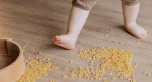IA barefoot toddler plays by stepping on dried macaroni that has spilled on the floor.