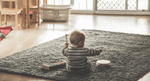 A baby playing on a grey rug in a minimalist room. The baby is playing with musical toys.