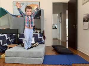 Image: A boy stands on a large cushion on the floor (Stuck Inside? Try These Gross Motor Games! Brittany Aquart Contributor Miami Mom Collective)