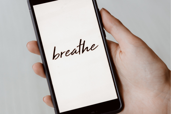 Image: A woman holding a phone with a screen that reads "breathe" (5 Tips to Manage Stress through Motherhood Zoe Costa Contributor Miami Mom Collective)