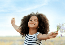 A young girl stands in a field with her arms spread wide, eyes closed and smiling. She is holding a bunch of wildflowers and the wind is blowing her hair.