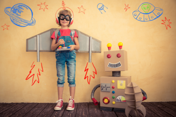 A child stands in front of a wall decorated like outer space. They are smiling, dressed in a homemade pilot’s helmet with goggles, cardboard rocket wings, and a joystick. A cardboard robot is standing next to them.