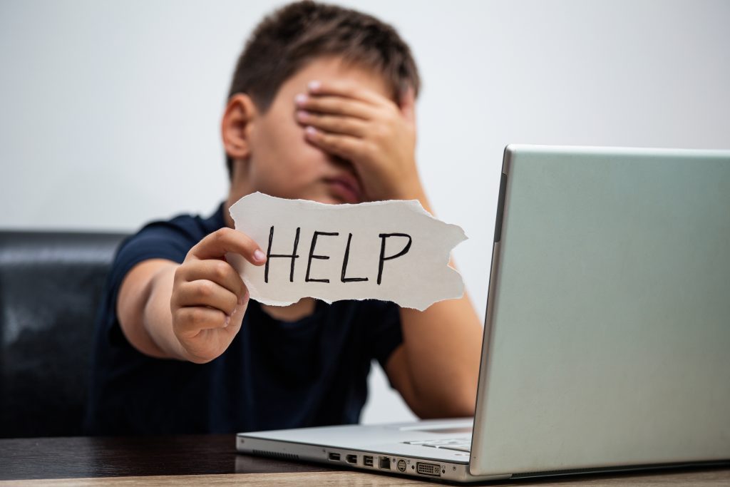 A boy sits in front of an open laptop holding out a strip of paper that has “HELP” written on it. He is covering his eyes with his hand and looks upset.