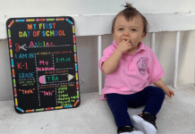 A toddler sits next to a My First Day of School board