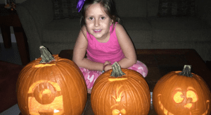 A little girl sits behind several creatively carved pumpkins