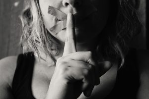 Image: A woman with tape over her mouth, holding up her finger like she's saying Shhhhh