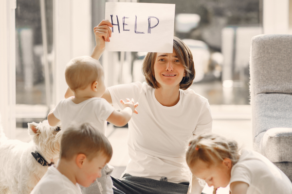 Image: A stressed out mom sits with her children and holds up a sign that reads, "HELP"